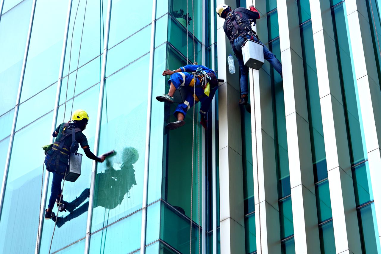 Trois laveurs de vitres suspendus à des cordes nettoient la façade vitrée d’un immeuble moderne en plein jour.